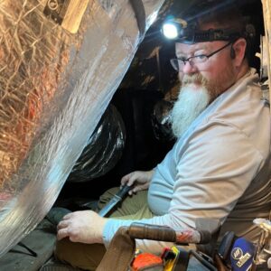 Rob Terry surrounded by duct work in an attic as he is maintaining a heat pump system